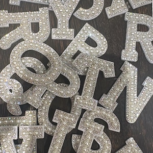 May include: A collection of white pearl-embellished letters on a dark wood background. The letters are made of white fabric and are covered in white pearls. The letters are in various sizes and are arranged in a random order.