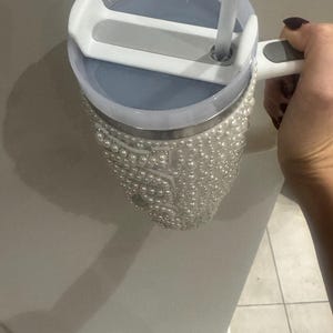 May include: A stainless steel tumbler with a pearl-covered exterior. The tumbler has a light blue lid with a white handle and a clear straw. The tumbler is held by a person.