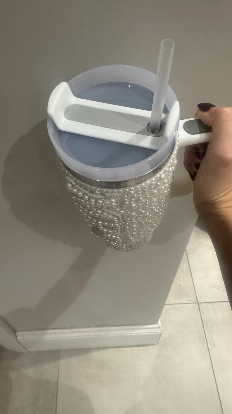 May include: A stainless steel tumbler with a pearl-covered exterior. The tumbler has a light blue lid with a white handle and a clear straw. The tumbler is held by a person.