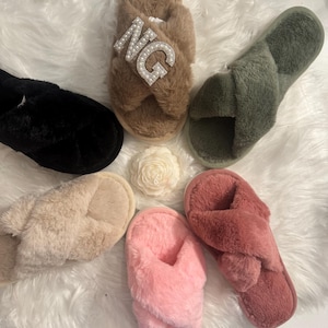May include: Six pairs of fuzzy slippers in various colors, including black, brown, beige, pink, and red. The brown slippers have a rhinestone embellishment that reads "NG".