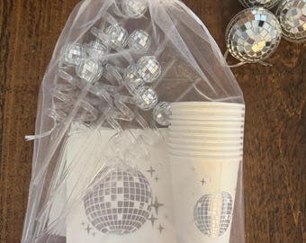 Op de afbeelding: Een witte organza tas gevuld met feestbenodigdheden, waaronder zilveren discobalbekers, rietjes en servetten. De tas bevat ook zilveren discobalversieringen.
