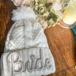 Op de afbeelding: Witte gebreide beanie met een nep bont pompon en het woord "Bride" gespeld in zilveren kralen.
