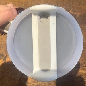 May include: A light blue, round lid with a white and gray handle. The lid has a gray, rectangular slider and a small, white, circular button. The lid is on a wooden surface.