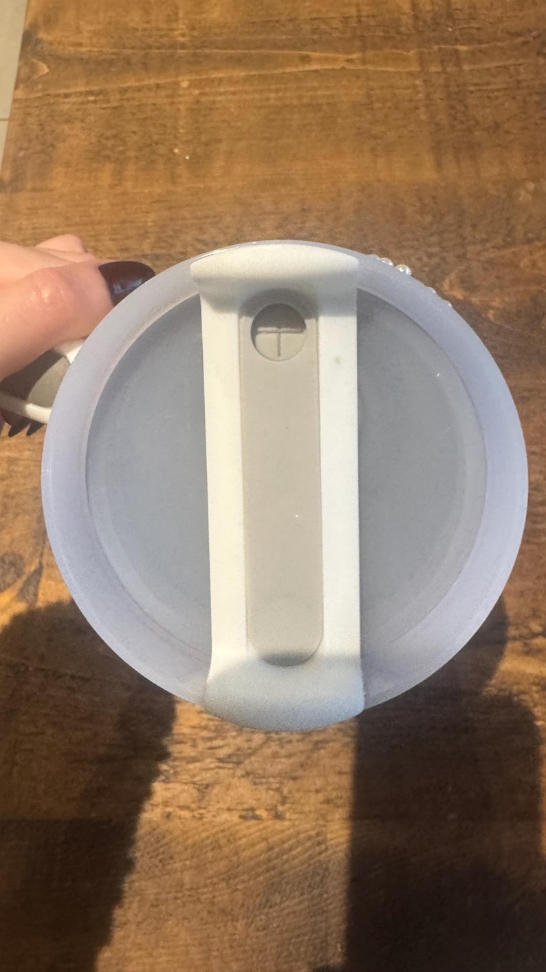 May include: A light blue, round lid with a white and gray handle. The lid has a gray, rectangular slider and a small, white, circular button. The lid is on a wooden surface.