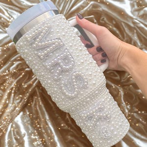 May include: White tumbler covered in faux pearls, with the word "MRS" spelled out in larger pearls. The tumbler has a white handle and a light blue lid with a straw. A heart-shaped embellishment is visible.