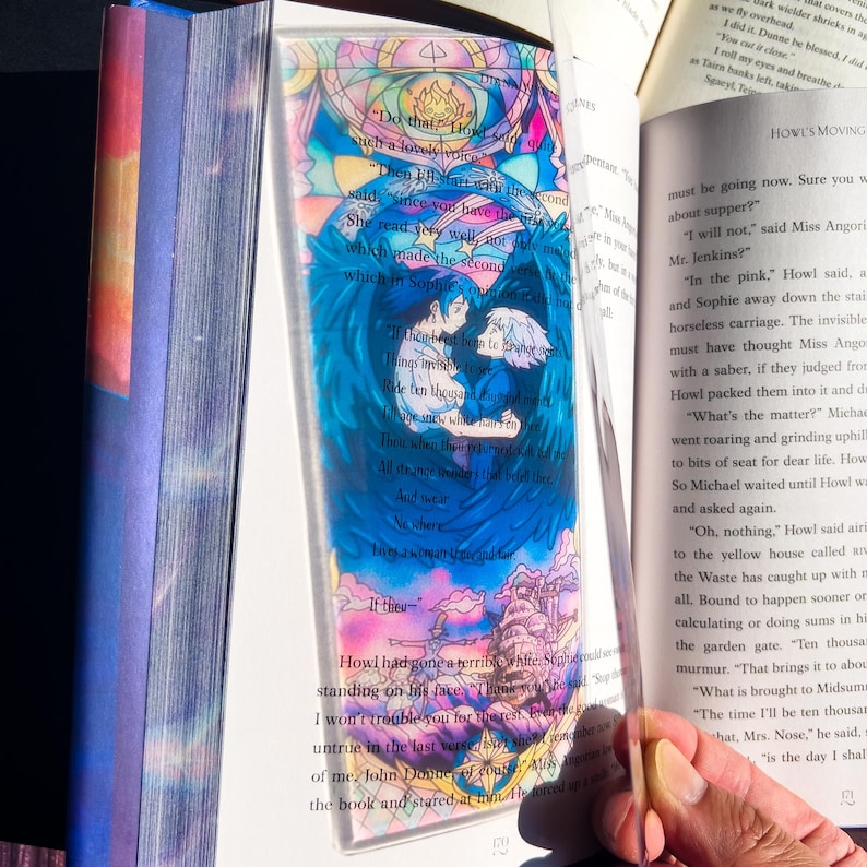 K&ouml;nnte beinhalten: Transparenter Acryl-Lesezeichen mit einer farbenfrohen Illustration von zwei Figuren mit blauen Fl&uuml;geln, vor einem Hintergrund im Buntglasstil. Das Lesezeichen enth&auml;lt Text aus dem Buch "Das wandelnde Schloss".