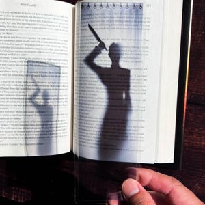May include: A close-up of a book with a shadow of a person holding a knife cast on the page. The shadow is dark and the knife is visible. The book is open and the pages are visible. The text on the page is blurry.