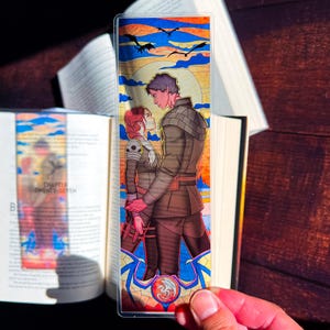 May include: A clear bookmark featuring a couple in a stained-glass style design. The artwork depicts a man and woman embracing, with a sunset and flying birds in the background. The bookmark is held in front of an open book.