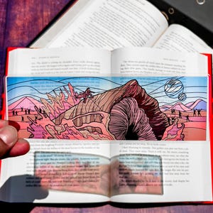 May include: A clear bookmark with a colorful illustration of a desert landscape with a large worm-like creature, a figure climbing a mountain, and silhouetted figures. The bookmark is placed on an open book.
