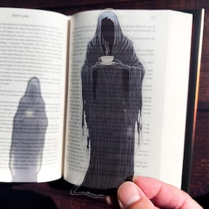 May include: A clear bookmark featuring a black and white illustration of a hooded figure holding a teacup. The figure is standing in front of a book.