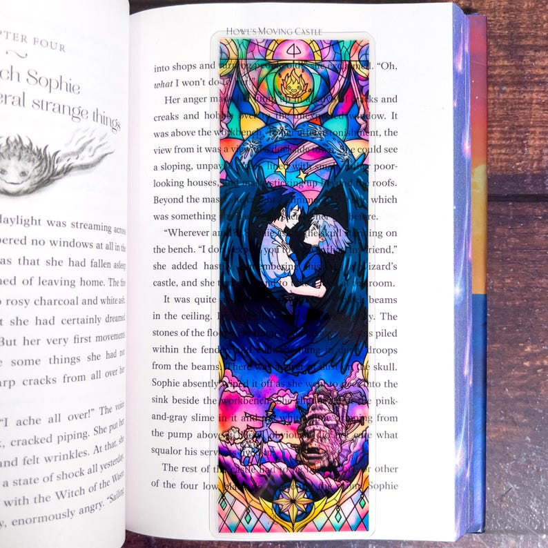 May include: A colorful bookmark featuring a stained-glass style illustration from Howl's Moving Castle. The design includes two characters embracing, surrounded by dark blue wings and a castle scene at the bottom. The bookmark is inserted in a book.