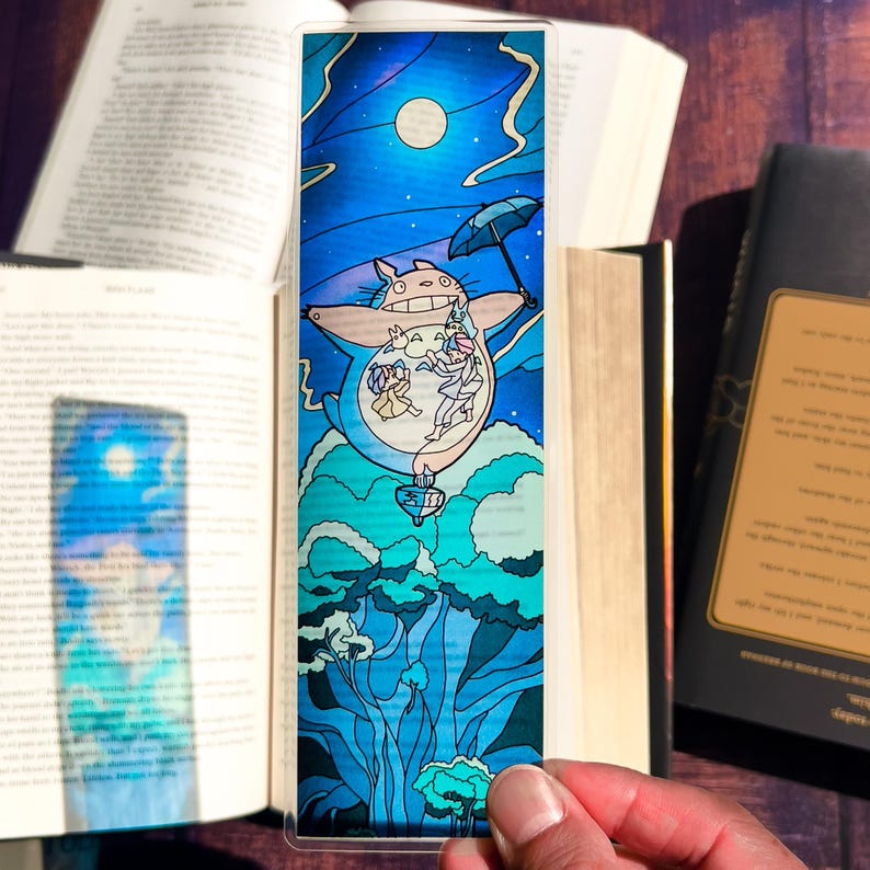 May include: A clear bookmark featuring a whimsical illustration of a large, blue, cartoon-like creature with an umbrella, surrounded by a night sky with a full moon. The creature appears to be carrying several smaller figures. The bottom of the bookmark shows stylized blue trees.