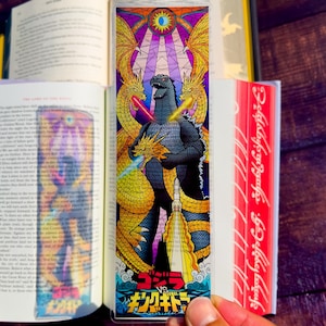May include: A rectangular bookmark featuring a stained-glass style illustration of Godzilla facing King Ghidorah. The design includes vibrant purple, yellow, and blue colors with Japanese text at the bottom reading Godzilla vs King Ghidorah.