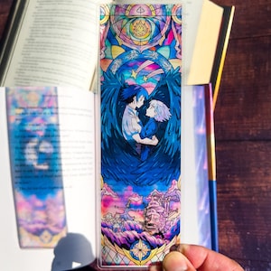 May include: A colorful bookmark featuring a stained-glass style illustration. The design depicts two figures embracing, surrounded by blue wings, stars, and a castle. The bookmark is rectangular and made of a clear, glossy material.