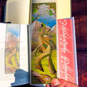 May include: A clear bookmark featuring a colorful illustration of a tree-like figure with a mountain backdrop. The design incorporates shades of green, brown, and yellow, with a blue sky and green foliage. The bookmark is held between the pages of a book.