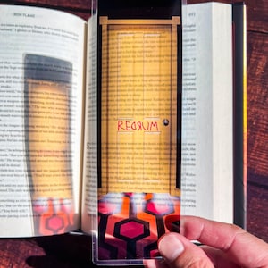 May include: A red and black patterned bookmark with a yellow door and the word "REDRUM" written in red. The bookmark is made of clear plastic and is designed to be used with a book.