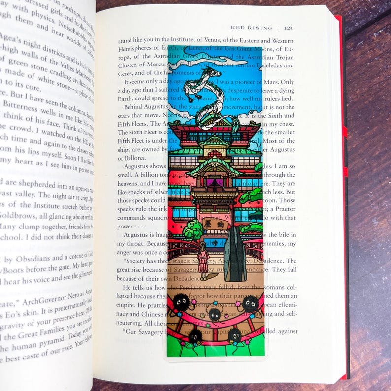 May include: A colorful bookmark featuring a Japanese-style building with a dragon and several small, black, round creatures. The bookmark is rectangular and is placed in an open book.