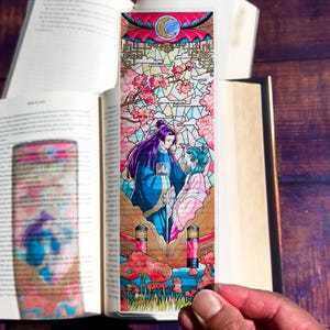 May include: A colorful bookmark featuring an anime-style illustration of two figures in traditional clothing. The design includes cherry blossoms, a moon, and a stained-glass effect. The bookmark is held in front of an open book.