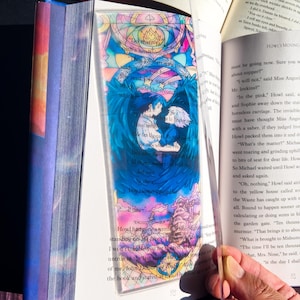 May include: A clear acrylic bookmark featuring a colorful illustration of two figures with blue wings, set against a stained-glass style background. The bookmark includes text from a book and is held open in a book.