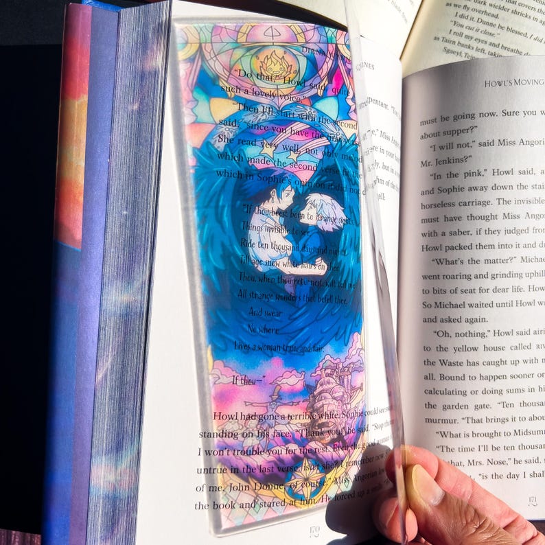May include: A clear acrylic bookmark featuring a colorful illustration of two figures with blue wings, set against a stained-glass style background. The bookmark includes text from a book and is held open in a book.