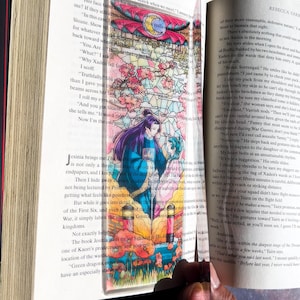 May include: A clear acrylic bookmark featuring a colorful stained-glass design with a fantasy illustration of two figures. The bookmark is inserted into a book with visible text.