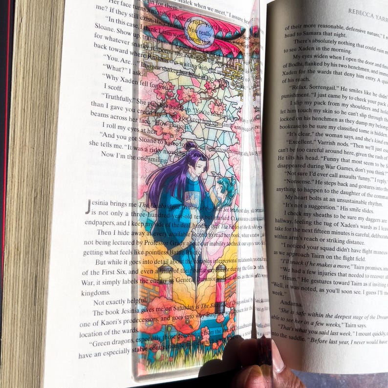May include: A clear acrylic bookmark featuring a colorful stained-glass design with a fantasy illustration of two figures. The bookmark is inserted into a book with visible text.