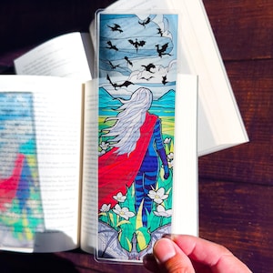 May include: A clear bookmark with a stained glass design featuring a woman with long white hair wearing a red cape and blue pants walking away from the viewer. The background features a field of flowers and a sky with flying black silhouettes of dragons.