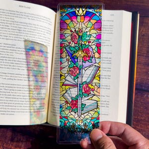 May include: A stained glass bookmark with a sword, roses, and books. The bookmark is colorful and features a rainbow of colors, including red, orange, yellow, green, blue, indigo, and violet. The bookmark is made of clear plastic and is designed to be used with a book.