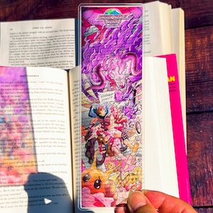 May include: A colorful bookmark featuring a fantasy illustration with the text "DUNGEON CRAWLER WORLD" and "GODDAMNIT DONUT!!". The artwork depicts characters and creatures in shades of purple, pink, and yellow.