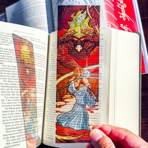 May include: A stained glass bookmark featuring a scene from The Lord of the Rings. The Balrog, a fiery demon, is depicted with a whip, facing off against Gandalf, a wizard with a white beard and a staff. The background is a fiery red and orange, with a bridge in the foreground.