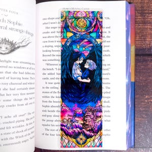 May include: A colorful bookmark featuring a stained-glass style illustration from Howl's Moving Castle. The design includes two characters embracing, surrounded by dark blue wings and a castle scene at the bottom. The bookmark is inserted in a book.