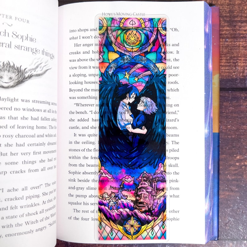 May include: A colorful bookmark featuring a stained-glass style illustration from Howl's Moving Castle. The design includes two characters embracing, surrounded by dark blue wings and a castle scene at the bottom. The bookmark is inserted in a book.