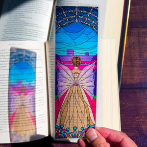 May include: A stained glass bookmark featuring a woman with wings standing in front of a castle. The bookmark has a pink, blue, and yellow color scheme.