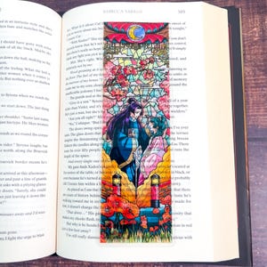 May include: A colorful bookmark featuring a stained-glass design with a romantic theme. The bookmark depicts two figures in ornate clothing, surrounded by flowers, a crescent moon, and a detailed architectural background. The bookmark is placed in an open book.