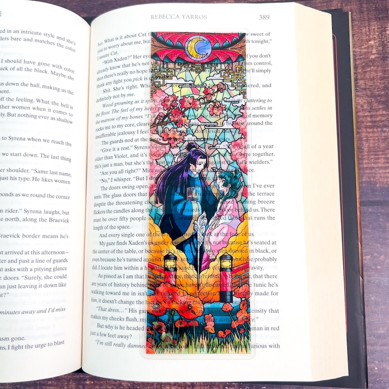 May include: A colorful bookmark featuring a stained-glass design with a romantic theme. The bookmark depicts two figures in ornate clothing, surrounded by flowers, a crescent moon, and a detailed architectural background. The bookmark is placed in an open book.