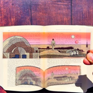 May include: A transparent bookmark featuring a desert landscape with a figure walking towards a structure. The bookmark has a pink and orange sky, with a dome-shaped building and other structures. The bookmark is placed on an open book.