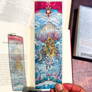 May include: A colorful bookmark with a stained-glass design. The artwork depicts a fantasy scene with figures, a castle, and wolves, set against a backdrop of blue and pink hues. A red bird-like figure is at the top.