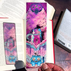 May include: A colorful bookmark featuring an illustrated scene with a woman and a man in a hat, set against a purple and pink background with lightning. A blue cat is at the bottom. The bookmark is held in front of an open book.