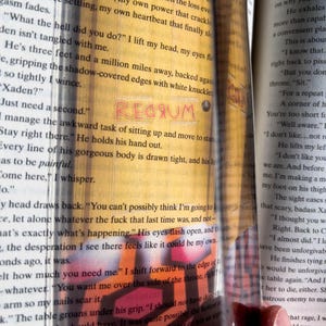 May include: Close-up of a book page with the word "REGRUM" written in red. The text on the page reads, "Just need a second." and "Stay right there." The book is open and the pages are yellowed.