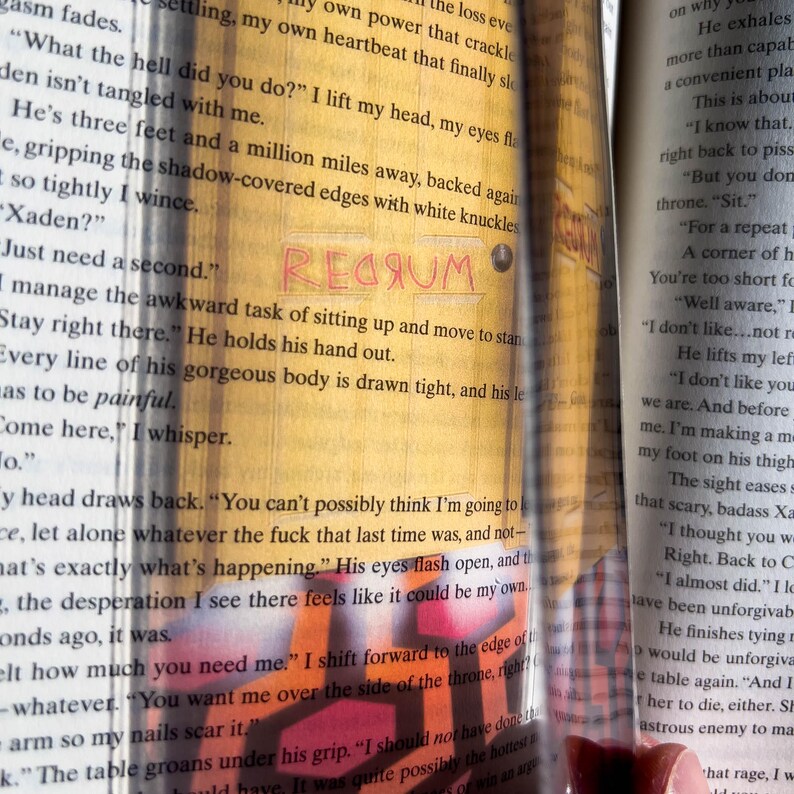 May include: Close-up of a book page with the word "REGRUM" written in red. The text on the page reads, "Just need a second." and "Stay right there." The book is open and the pages are yellowed.