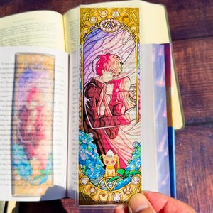 May include: A colorful bookmark featuring a romantic anime-style illustration of a couple embracing. The design includes a stained-glass effect with gold accents, a cat character, and various other character icons. The bookmark is held open in a book.