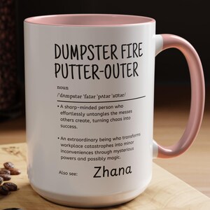 May include: White ceramic mug with a pink interior and handle. The mug features the text "DUMPSTER FIRE PUTTER-OUTER" with a definition and the name "Zhana". A spoon, coffee beans, and a pastry are on a wooden surface.