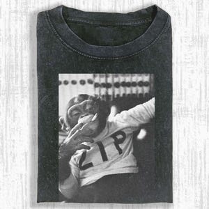 Puede incluir: Camiseta gris oscuro con una imagen en blanco y negro de un chimpancé que lleva una camisa blanca con la palabra "ZIP" impresa. El chimpancé se está llevando algo a la boca. La camiseta está doblada.