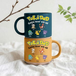 Könnte beinhalten: Zwei Keramikbecher, die übereinander gestapelt sind. Der obere Becher ist teal, der untere senfgelb. Beide Becher tragen den Text "PokéDad Gotta love' em all" mit Illustrationen von Zeichentrickfiguren und Namen. Die Becher haben einen Henkel.