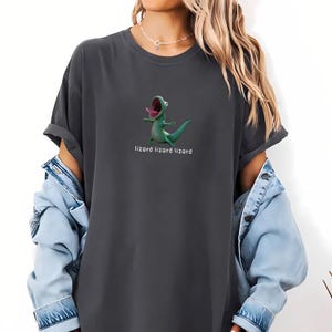Puede incluir: Camiseta gris oscuro con un dibujo de un lagarto verde de dibujos animados, con la boca abierta y la lengua fuera. El texto "lizard lizard lizard" está impreso debajo. La camiseta tiene mangas enrolladas.