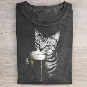 以下が含まれることがあります： ビールグラスからビールを飲む縞模様の猫の白黒画像が特徴の濃い灰色のTシャツ。猫の顔が見え、足でグラスを持っています。シャツは明るい木製の表面に折りたたまれています。