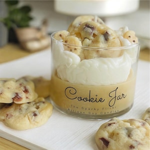 May include: A clear glass jar candle with a cookie theme. The candle has a light brown base, a white layer, and cookie-shaped wax pieces on top. The jar is labeled "Cookie Jar" with "The Sparky Flicker Artisan Soy Candles" below.
