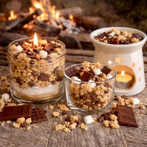 May include: A lit candle in a glass jar filled with s'mores ingredients, including chocolate chunks, marshmallows, and graham cracker crumbs. A small glass and a ceramic warmer with similar ingredients are also present, along with chocolate bars.