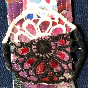 May include: A close-up of a handmade fabric embellishment with a black lace design, red glass beads, and a white lace overlay. The embellishment is attached to a purple and pink fabric background.