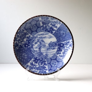 May include: A blue and white ceramic plate with a Japanese-inspired design. The plate features a landscape scene with trees, a river, and a house. The rim of the plate is decorated with a brown band.